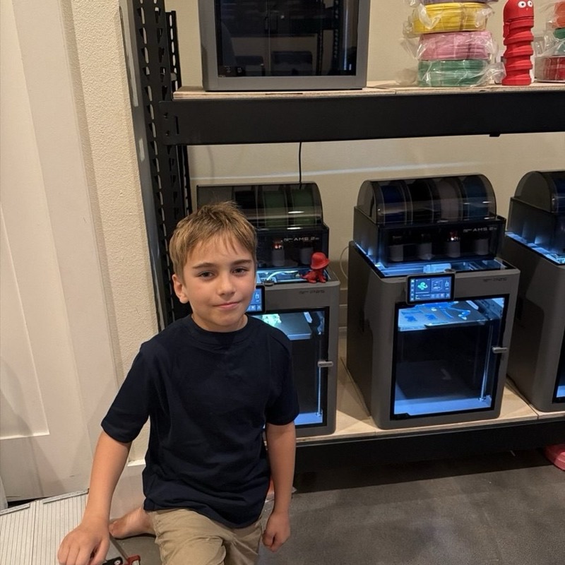 A shelf of enclosed Bambu Lab 3D printers at the Printer Ranch in Austin, with Joseph kneeling in front
